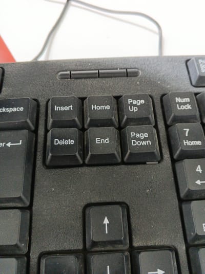 A close-up, eye-level shot shows the top section of a black computer keyboard, with a white background and a gray cable visible in the upper portion of the frame. The keyboard itself appears dusty. Prominent keys include "Backspace," "Insert," "Home," "Page Up," "Page Down," "Delete," "End," "Num Lock," and the numeric keys "7," "4," and a partial view of "1." An upward-pointing arrow key is also visible in the bottom section of the frame. The image is of a keyboard and does not depict any scene in the city of Hulbuk, Tajikistan.