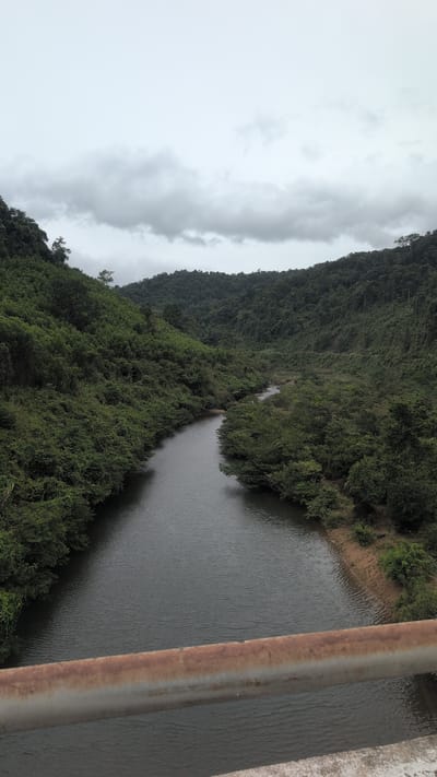 The image shows a serene river flowing through a lush green valley. Dense foliage covers the rolling hills on either side, with a cloudy sky overhead. In the foreground, a rusty metal railing runs horizontally across the frame, suggesting the viewpoint is from a bridge. The river, with its dark, rippling water, winds its way into the distance, disappearing among the trees. The scene evokes a sense of natural tranquility, likely captured in a rural or mountainous area. The location is identified as Trường Sơn Commune, Vietnam.