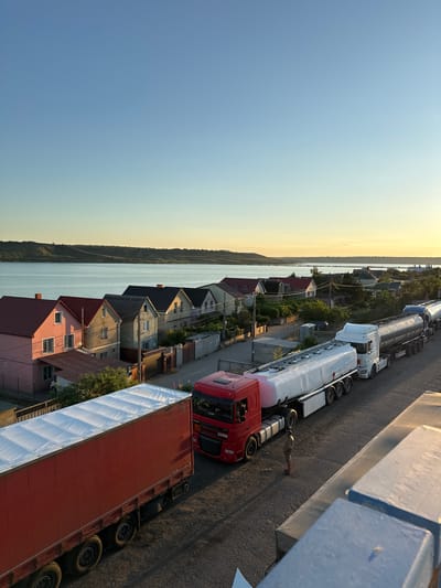 The image captures a street scene in Yelyzavetivka, Ukraine, at golden hour. The foreground features the back of a red semi-trailer truck, its cargo covered with a white tarp. Adjacent to it, a line of white semi-trailer tanker trucks is parked. A lone individual stands on the asphalt between the red truck and the tanker trucks. In the background, a row of houses with red and dark roofs lines a street. Beyond the houses, a body of water, likely a river or reservoir, stretches across the frame, with a grassy hillside visible on the far bank. The sky is clear and displays a gradient of blue and yellow hues, indicative of sunset.