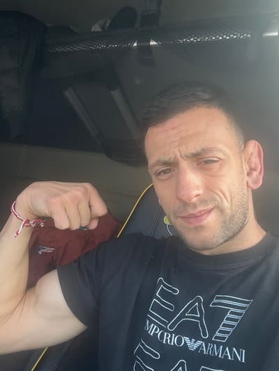 A close-up selfie depicts an adult male seated within an interior space, likely the cabin of a vehicle. The male, with short dark hair and facial stubble, wears a black t-shirt featuring the white "EA7 EMPORIO ARMANI" logo. A red and white braided string bracelet is visible on his left wrist. He is flexing his left bicep, with his left arm bent and fist clenched, while looking directly at the camera with a slight smirk and furrowed brow. Behind him, a dark seat with yellow piping is partially visible. Above his head, an overhead compartment with black mesh netting contains dark objects or luggage. The environment's specific location within the city of Tortoreto, Italy, cannot be determined from the visual evidence provided.