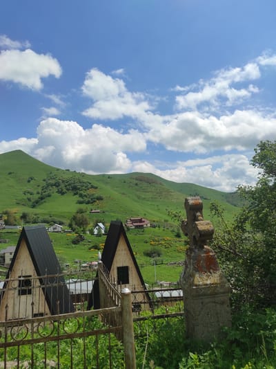 The image depicts a bright, sunny day in Kumlistsikhe, Georgia, with a clear blue sky dotted with fluffy white clouds. In the foreground, a weathered stone cross stands sentinel, partially obscured by lush green foliage and a rusty metal fence. Beyond the fence, two triangular structures with dark roofs are visible, hinting at the presence of houses or other buildings. The middle ground is a vibrant expanse of rolling green hills, speckled with yellow wildflowers and scattered with more houses and buildings that form a small village. The backdrop is dominated by the majestic, verdant mountains characteristic of the Georgian landscape. The overall scene evokes a sense of rural tranquility and natural beauty.