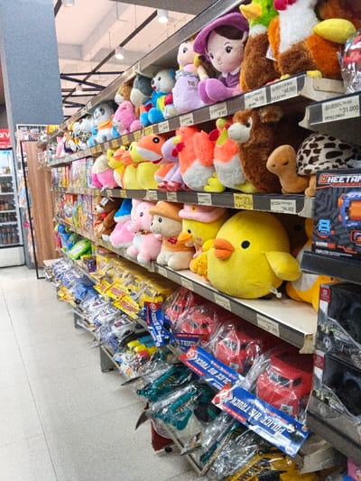 The image captures a brightly lit toy section within a retail store, likely a supermarket or department store, located in Sindanglaya, Indonesia. The perspective is slightly elevated, looking down an aisle packed with merchandise.
The upper shelves are densely arranged with a colorful assortment of plush toys. These include large yellow stuffed chicks or ducks with orange beaks, pink elephant-like figures, brown bears, human-like dolls dressed in purple, orange foxes, and a distinctive brown and white spotted turtle. Many of these toys have price tags attached, with visible amounts such as "48.000," "54.000," and "31.000," indicating prices in Indonesian Rupiah. Other plush toys visible include blue and white pandas, white and orange roosters, and various other animal shapes in vibrant hues.
The lower shelves display packaged toys and other items, predominantly wrapped in clear plastic. Prominently featured are multiple "POLICE BIG TRUCK" toy sets, some in blue and others in red packaging. Other packaged items include small red truck toys and other plastic vehicles. Text labels like "TRUCK," "OLICE BIG TRUCK," and "POLICE BIS TRUCK" are visible on these packages. Additional text from product labels includes "CHEETAN" and "TORTOARE." On the far left, a vertical sign partially visible displays the text "arv," possibly part of the store's branding.
The scene suggests an active retail environment, with products neatly displayed for sale. No people are visible, emphasizing the merchandise and the organization of the store's toy department. The lighting appears to be artificial, typical of an indoor commercial setting.