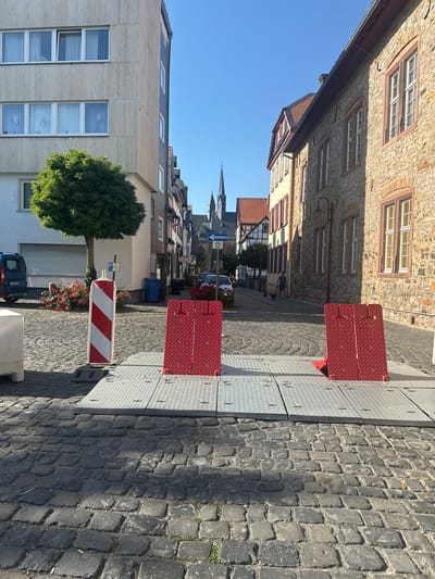 The image shows a street scene in Butzbach, Germany. The perspective is from ground level, looking down a cobblestone street lined with historic stone buildings on the left and metal construction plating on the right. The sky is clear and blue, suggesting a sunny day. The time of day appears to be late morning or early afternoon, given the shadows.

On the left side of the street, there are several multi-story buildings with stone facades, featuring numerous windows with reddish-brown frames. Some windows have white shutters. Further down the street, the architecture changes to half-timbered buildings. A large, lush green tree is prominent in the mid-ground.

On the right side of the street, metal plates are laid out, likely for construction or temporary access, with red metal barriers placed on top. A striped red and white barrier pole is also visible.

In the distance, a church spire can be seen above the rooftops, indicating the presence of a town center or historic district. A blue van is parked on the left side of the street.

Visible text includes a parking sign with a "P" symbol and a blue road sign with a bicycle symbol and German text, possibly indicating a bike path or route. Another smaller white sign with an inscription is also present. There is no discernible activity or event taking place beyond the ordinary street view.