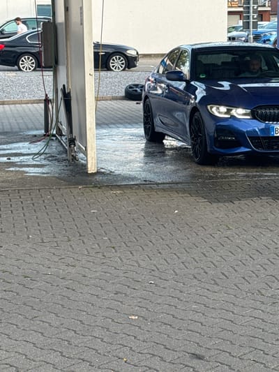 The image depicts a street scene, likely in Ahlen, Germany, featuring a blue BMW sedan being washed at a self-service car wash. A black BMW sedan is parked to the left, with a person wearing a white shirt standing near it. The ground is paved with interlocking concrete blocks, and water from the car wash has created wet patches. In the background, other vehicles and buildings are visible, suggesting an urban setting. The blue BMW is the primary subject, showcasing its headlights and front grille. The presence of car wash equipment, like hoses and a water spraying apparatus, indicates the ongoing activity.