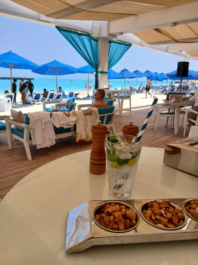 A photograph captures a scene along the coast of Sidi Abdel Rahman, Egypt. The foreground features a round table where a tall glass filled with ice, lime, and a blue and white striped straw rests, alongside two wooden salt and pepper grinders and a silver tray holding three metal bowls filled with peanuts. In the background, there is a row of blue sun umbrellas and white beach chairs lining the shoreline. People are seen lounging under the umbrellas. The sky is clear and the water is light blue, typical of a coastal setting. The photo appears to be taken from under a shaded area with tan-colored awnings and supporting beams.