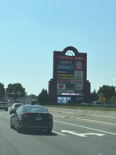 The image depicts a highway scene in Fredericksburg, Virginia, USA, on a clear, sunny day. The focal point is a tall, multi-level signpost for "CENTRAL PARK," a shopping center. The sign displays the logos and names of various stores, including Lowe's, Walmart, Target, Kohl's, Sephora, Best Buy, Ashley HomeStore, Hobby Lobby, and T.J. Maxx, among others. At the bottom of the signpost, a digital display reads "FUN FOR ALL AGES."
Several vehicles are visible on the highway. In the foreground, a black Honda Civic is driving in the right lane. Behind it, a white SUV and a white truck are also present. The highway has multiple lanes, separated by concrete barriers and white lane markings. Traffic lights and trees line the side of the road. The sky is a clear blue.