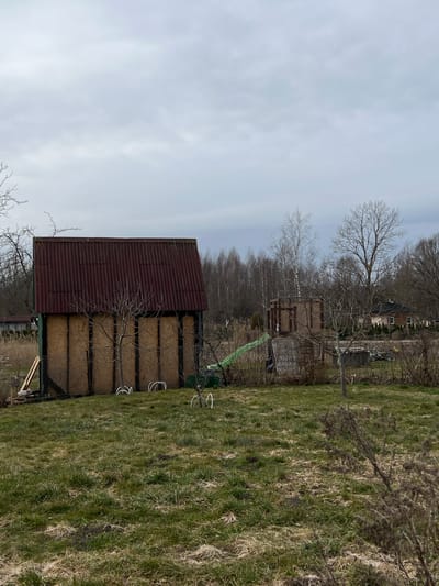 The image shows a corrugated metal structure, possibly a barn or shed, located in a grassy area with trees in the background. The sky is overcast. Based on the presence of the place in the prompt, the location is likely in Dārzciems, Latvia. No people, animals, or interactions are visible in the frame.