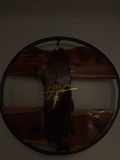 In Miami Shores, United States of America, a circular, custom-crafted wall clock is observed hanging on a light-colored, dimly lit interior wall. The clock, approximately 2-3 feet in diameter, features a dark, likely metal, outer frame. Within this frame, three horizontal segments of natural-edge, reddish-brown wood, possibly cedar, are arranged and coated with a clear, glossy resin, creating a reflective surface. A prominent, irregular, vertically oriented piece of dark, highly polished material, possibly dark wood or resin, overlays the center of these horizontal segments. This central piece exhibits organic contours, including a jagged top edge. Gold-toned clock hands, one ornate and the other simple, are centrally mounted on this dark element, indicating approximately 9:15. A dark, thin hanging apparatus is visible at the top center of the clock's frame. No individuals or dynamic actions are present in the scene, which appears to be a static interior setting.