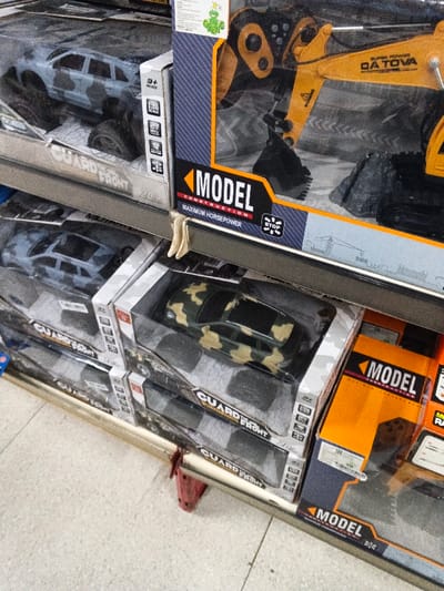 A shelf displays toys in their packaging. On the top shelf, a gray and blue camouflage toy car sits beside a yellow excavator. The excavator's packaging emphasizes "Model Construction" and "Maximum Horsepower." Below, rows of the same toy car in gray camouflage and green camouflage are stacked, labeled "Guard The Front." Next to the green camouflage cars, an orange toy is visible, also labeled "Model."