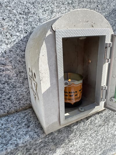The image displays a close-up, angled view of a light-colored stone or concrete memorial shrine attached to a speckled gray and white granite wall or monument in Nof HaGalil, Israel. The shrine features a carved Star of David on its left exterior surface. A metallic door, hinged on the right and adorned with a brick-like pattern around its frame, is open, revealing the interior cavity. Inside, a cylindrical metallic memorial candle, featuring an orange and black label with Hebrew text and "MEMORIAL CANDLE," is actively burning, indicated by a visible flame. Direct sunlight illuminates the scene, casting a distinct shadow of the shrine onto the granite surface. No individuals or direct interactions are visible.