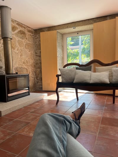The image depicts the interior of a room located in Oliveira de Frades, Portugal. A seated individual's lower legs and feet are visible in the foreground, extended towards the viewer. The person wears light blue denim jeans and dark brown pointed-toe flat shoes with an ankle strap. The floor is covered in terracotta-colored square tiles, showing variations in tone.

To the left, a modern black-framed wood-burning stove with a glass door rests on a light grey stone or concrete hearth. A stainless steel flue extends vertically from the stove, ascending against a wall constructed from irregularly shaped, light-colored rough-hewn stones. This stone wall continues across the back of the room.

Centered in the background is a white-framed window, through which lush green foliage, possibly trees or bushes, and a glimpse of a light-colored building are visible outdoors. The window is flanked by two tall, light-colored wooden panels, possibly doors or custom cabinetry.

To the right of the window, a dark-stained wooden sofa with a caned back and dark upholstered seat is positioned. Multiple cushions are arranged on the sofa: predominantly light-colored (off-white/light grey) square and rectangular pillows, with one darker grey cushion visible underneath. Natural sunlight streams through the window, casting bright patches onto the tiled floor. The ceiling is plain white.