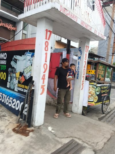 A man wearing a black shirt and a baseball cap is holding a baby in his arms, standing between two white pillars adorned with orange numbers reading "1781945" and "1782025," likely dates or commemorations. A food cart is on the right, displaying "MIE KOCOK" and Indonesian text. Behind the man is a red wall. A sign with promotional text and a phone number (531652002) is partially visible on the left. Above them is a white beam with the inscription "DIRGAHAYU INDONESIA KE-80 TAHUN."