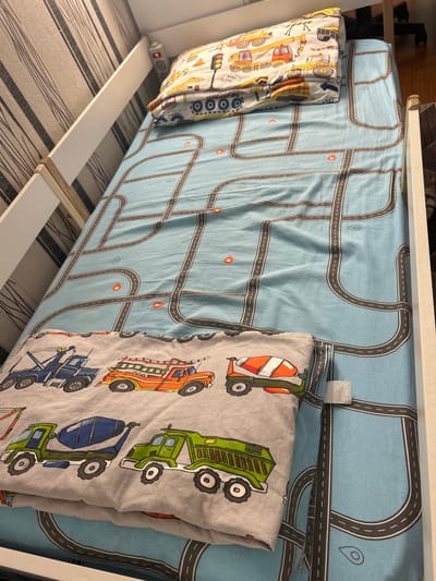 This is a close-up, high-angle shot of a child's bed with bedding featuring construction vehicles and a road map pattern. The bed appears to be a loft bed, with a white wooden frame visible on the left and right sides. The bedding includes a light blue sheet with a dark gray road pattern and orange traffic cones. A folded blanket or duvet with a gray background and colorful illustrations of construction vehicles (tow truck, fire truck, cement mixer, dump truck) is placed on top of the sheet. A pillow with a similar construction vehicle pattern is at the head of the bed. The background wallpaper has a striped pattern in shades of gray and white. A white electrical outlet strip with multiple sockets is attached to the white frame on the left side of the bed, near the pillow. The lighting suggests an indoor environment, possibly a bedroom. The image does not provide any specific information about the city of Smolyaninovo, Russia.