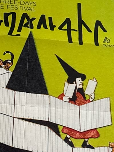 This image is a close-up of a poster with a lime green background. In the upper left corner, white text reads "THREE-DAYS" and "E FESTIVAL". In the center of the poster, a stylized black cat with a white spot on its chest is depicted looking to the left. Below and to the right of the cat, a witch wearing a black pointed hat, a red patterned dress, and black shoes is reading two books. The witch is positioned behind a white structure that resembles stacked books or shelves, and she appears to be sitting. To the right of the witch, a signature reads "MAY" with the numbers "15,16,17" underneath. In the lower right corner, a brown and white cat is visible. The overall aesthetic suggests a whimsical and festive event. The location in the city of Jrarat, Armenia is not specified within the image.