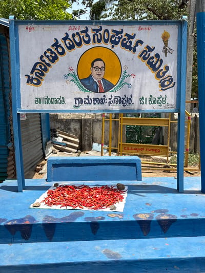 The image shows a scene in Chikkarampur, India. A sign in Kannada script is prominently displayed, bearing the name "Karnataka Dalit Sangharsh Samiti," a Dalit organization. The sign also features a portrait of B. R. Ambedkar, a key figure in the Dalit movement. Below, the sign specifies "Taaluk: Gangawati, Grama Shakha: Sanapur," indicating the regional and local branch details. To the right, dried red chilies are laid out on a white cloth, placed on blue concrete steps. This setup is partially shaded, casting distinct shadows on the blue surface.
