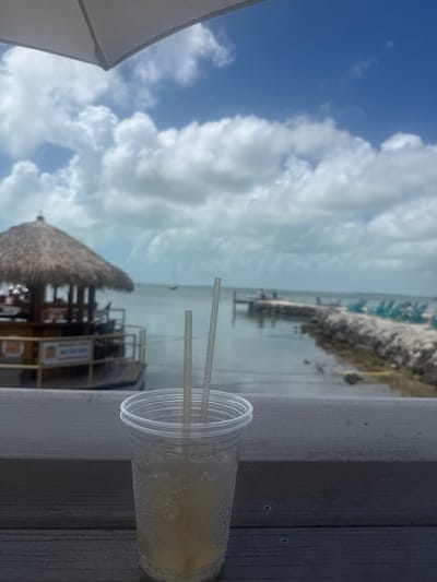 The image captures a view from what appears to be a deck or ledge in Key Largo, Florida, USA. In the foreground, a clear plastic cup with two straws sits on a grey wooden surface. The cup contains a light-colored liquid with visible condensation on the exterior. In the middle ground, a thatched-roof structure, possibly a bar or kiosk, is visible. Behind it, there is a body of water, likely the ocean, extending to the horizon. A pier or jetty extends into the water with some structures on it, and further out are hints of an island. The sky is partly cloudy with patches of blue visible. A white umbrella is partially visible in the bottom left corner of the image.