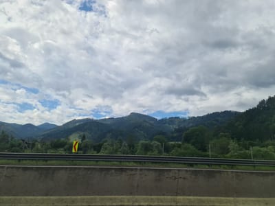 The image depicts a scenic view of a mountainous landscape, likely from a road in or near Brunn, Austria, given the context provided. The foreground shows a concrete barrier, likely part of a highway or road infrastructure, with a metal guardrail above it. Beyond the barrier, a vibrant green forest covers the rolling hills and mountains that dominate the midground and background. The mountains are densely covered with trees, creating a textured green expanse that stretches towards the horizon. Patches of blue sky peek through a dramatic sky filled with large, white, and gray clouds, suggesting a dynamic weather system. A yellow and red directional traffic sign, indicating a curve, is visible on the left side of the road, further suggesting a road setting. No people or animals are present in the image. The overall impression is one of natural beauty and a sense of travel along a scenic route.