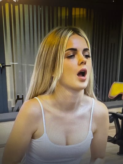 The media file shows a close-up, horizontally oriented, full-body shot of a young, fair-skinned person with blonde hair. They are wearing a white tank top. The person's mouth is open, and their eyes are looking upwards. The background appears to be an indoor setting with horizontal lines and some dimly lit objects, possibly gym equipment. There is a hint of a yellow shape above and behind the person's head. The lighting suggests it could be artificial light, and there are no clear indicators of the time of day or weather. The emotional expression of the person is one of surprise or exertion. There is no visible text in the image. The context of Chorzów, Poland is provided, but there are no specific visual cues within the image to confirm this location.