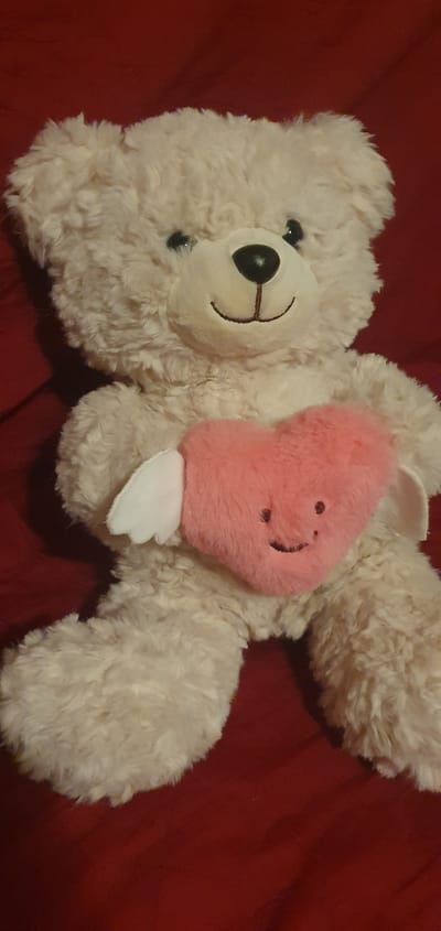 A plush teddy bear with cream-colored fur is positioned centrally against a dark red background. The bear has black button eyes, a black nose, and a stitched smile. It is holding a pink, heart-shaped pillow with a simple embroidered smiley face. The pillow has two small white wings attached to its sides. The bear's paws are resting on its lap, one slightly overlapping the heart pillow. The lighting appears soft, casting subtle shadows. There is no information about the city of Alūksne, Latvia, present in the image.