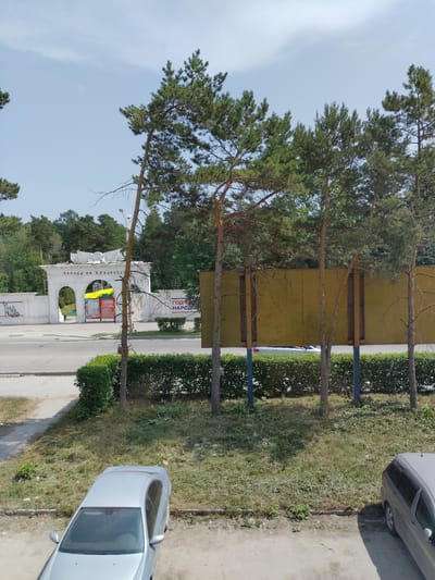 The image depicts a street scene in Iskitim, Russia, bathed in the bright light of a sunny day. Tall pine trees with dense green foliage line the left side of the frame, casting shadows onto a paved area and a grassy verge. A white, ornate archway stands prominently in the background, with arched openings and decorative elements. On the archway, text is visible, appearing to read "ГОРОД ИМ. Н. И КОРОТЕЕ" and below it "СЛАВЫ". Above the arch, a yellow and green flag is displayed, possibly the flag of the region or a specific event. Several posters or banners are also attached to the archway, some featuring text and images.

Beyond the archway, a street stretches into the distance, bordered by more greenery and what appears to be a building with a large yellow facade. To the right of the street, a neatly trimmed hedge runs along the property line. A silver car is parked on the right side of the image, its rear end visible. Another silver car is partially visible in the upper right corner of the frame. The overall scene suggests a public space, possibly an entrance to a park or town square, decorated for an event or celebration. The clear sky and strong sunlight indicate it is daytime, likely late morning or afternoon.