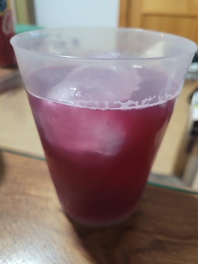 A close-up, slightly angled shot shows a clear plastic cup filled with a reddish-purple liquid and ice cubes. The cup has a frosted texture and a slightly flared rim. The liquid has a foamy layer near the top. The cup is placed on a wooden surface. In the background, out of focus, a red object with some text and a green object are visible. The environment appears to be indoors, possibly a kitchen or dining area, with wooden doors and walls in the background. The image does not provide any specific visual cues or context that would allow for the determination of its location in Els Campellos, Spain.