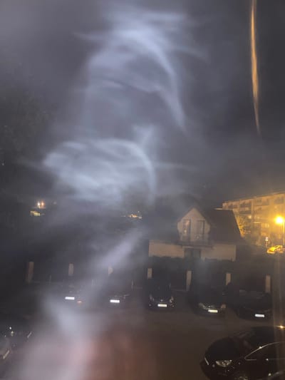 A night photograph taken from an elevated perspective in Konstantynów Łódzki, Poland, shows a dense fog or smoke obscuring much of the scene. A residential building with a dark roof and pale walls is visible, with lights on inside. In front of the building, several parked cars are dimly illuminated, some with visible license plates. Further in the background, a multi-story apartment building is lit with warm yellow lights. The sky above is dark and appears to have swirling patterns of light or thicker fog, possibly illuminated by streetlights or other urban light sources. A tall, thin, bright yellow light streak is present on the far right side of the frame, possibly from a streetlight or a long exposure effect. The foreground is largely obscured by the fog, with the tops of dark-colored vehicles visible. The overall atmosphere is hazy and somewhat mysterious due to the heavy fog.