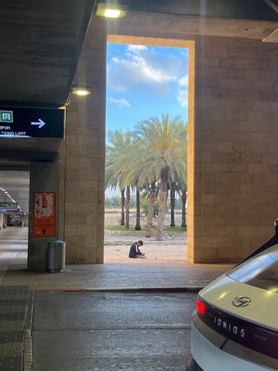 The image shows a view from inside a covered area with an open doorway. The doorway is framed by stone walls and leads to a courtyard with a few palm trees. The courtyard is empty except for a person sitting on the ground. In the foreground, a white Hyundai Ioniq 5 is parked. The person in the courtyard appears to be looking down at the ground and may be cleaning something or looking for something. The covered area contains a sign in Hebrew and English that reads "Parking Laot." There is also a trash can and an orange poster with arrows pointing to the right and up. There are lights on the ceiling of the covered area. The sky is bright blue with some white clouds. The image is likely taken at Ben Gurion Airport, Israel.