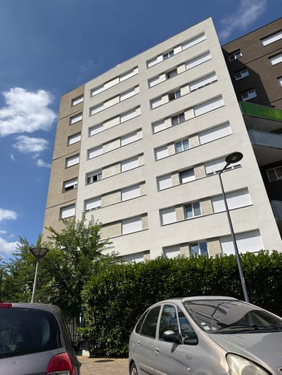 This is a low-angle, full shot of modern apartment buildings in Évry-Courcouronnes, France, under a bright blue sky with scattered clouds. The architecture is characterized by clean lines, alternating light and dark grey facades, and numerous elongated windows with white slatted blinds. The buildings appear to be tall and multi-story.
In the foreground, a silver car is parked on a paved area, partially obscured by a dense green hedge. Further back, a dark grey car is also visible, facing away from the camera. A streetlamp with a triangular head is positioned between the buildings and the cars, extending outwards.
The scene suggests a sunny day, likely late morning or early afternoon, given the strong shadows cast by the buildings. The overall impression is one of urban residential living, with a focus on the architectural design and the interplay of light and shadow. There is no discernible activity or event taking place, and no visible text. The image is captured with a slightly tilted perspective, adding a dynamic feel to the composition.