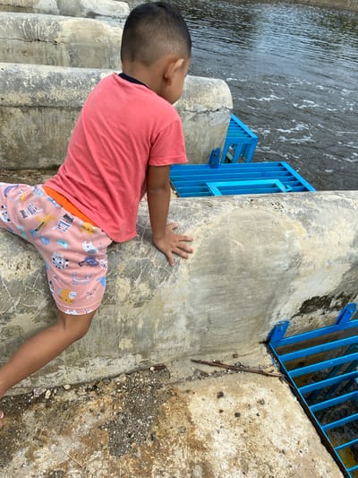 A young boy wearing a coral pink t-shirt and patterned shorts is positioned on a concrete structure near a body of water. The boy is facing away from the camera and towards the water, with his left arm resting on the concrete and his right hand reaching forward. The concrete structure appears to be made of stacked, curved sections, possibly part of a dam or weir. Adjacent to the boy is a bright blue metal grate structure, which sits over the water. The water itself is choppy, indicating some current or movement. The ground beneath the concrete structure is rough, with small stones and dirt visible. The environment suggests an outdoor, possibly recreational or functional area, in Aneuk Paya, Indonesia.