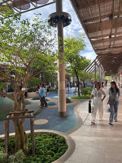 In Krabi, Thailand, an outdoor walkway covered by a wooden lattice roof leads to a vibrant public space. Two young women are walking in the foreground. One, in a grey top and jeans, has a small crossbody bag and is looking down. The other, in a white outfit, poses with her hand on her hip. Beyond them, the walkway opens into a park-like area with a colorful, multi-textured floor featuring blue and green abstract shapes. A large, textured pillar, possibly a climbing structure for cats, stands prominently. Lush green trees and bushes provide shade. In the background, other people are visible, some sitting, others standing, suggesting a leisurely day. The sky is partly cloudy, indicating daytime. The overall scene conveys a relaxed and welcoming atmosphere.
