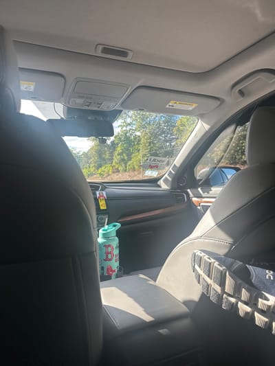 The image displays the interior of a vehicle, viewed from a rear passenger seat looking forward towards the dashboard and windshield. The vehicle is situated in Newton, United States.
Inside the vehicle, the dark-colored backs of the driver and front passenger seats are prominent. A light teal water bottle, featuring a red "B" logo and "BOS RED SOX" text, is positioned in a cup holder on the center console. A dark blue athletic shoe with a distinct textured sole rests on the lower part of the front passenger seat. The dashboard is dark with a horizontal trim. A rearview mirror is mounted centrally, with a dashcam or similar recording device attached beneath it. Uber and Lyft decals, along with a yellow human-figure sticker, are affixed to the windshield and dashboard, indicating the vehicle is operating as a ride-sharing service. Sun visors are present, with a warning label visible on the passenger side.
Outside the vehicle, seen through the windshield, is an environment dominated by green trees and foliage under a bright blue sky with sparse clouds. A dark road surface and a strip of bare earth are visible immediately beyond the vehicle. A portion of another dark-colored vehicle is visible through the front passenger window.