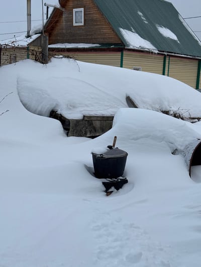 This image depicts a deeply snow-covered outdoor scene in Probuzhdenie, Russia, during winter. The background features a two-story house: the upper level is clad in light brown wooden siding with one visible white-framed window, while the lower level features lighter-colored horizontal siding with dark green trim and a smaller, partially obscured window. The house is topped by a steeply pitched dark green corrugated metal roof, with patches of snow and several icicles hanging from the eaves. A chain-link or wire fence is partially visible on the far left, mostly covered by snowdrifts.
The foreground and midground are dominated by extensive, untouched white snow, forming deep, undulating drifts that obscure the underlying ground and structures. In the central foreground, a dark, lidded pot or bucket, possibly cast iron, is positioned on a partially buried, multi-legged dark metal stand. A wooden-handled utensil, appearing to be a ladle or spoon, protrudes vertically from the pot's lid. To the immediate right of the pot, a large, dark, cylindrical object, consistent with a section of pipe, lies partially buried in the snow. Faint footprints are visible in the fresh snow in the lower-left foreground, indicating recent passage. No people are present in the scene.