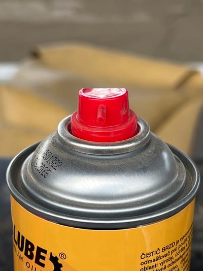 The image shows a close-up of the top of a yellow spray can with a red nozzle. The can has some text printed on it, partially visible as "LUBE" and other text in what appears to be Czech. There is also a small registered trademark symbol. The can is slightly dusty. The background is blurred and appears to be outdoors, with some earthy tones and shapes that could be natural elements or structures. There is no information about the location in Lanžhot, Czechia.