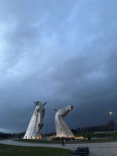 The image depicts The Kelpies, two large metallic horse head sculptures, located in Helix Park, Falkirk, United Kingdom, under a dark, heavily overcast sky indicative of dusk or early evening.
The two prominent equine sculptures dominate the middle ground. The left sculpture faces primarily left and slightly forward, its head angled downward. The right sculpture faces right, its head tilted upwards with its mouth slightly ajar. Both sculptures are constructed from numerous interlocking metallic plates, giving them a textured, segmented appearance, and are illuminated from their bases by warm yellow uplighting. They stand within a circular water feature or shallow pool.
Surrounding the sculptures are paved pathways and green grassy areas. Several small, indistinct human figures are visible on the pathways near the base of the sculptures, appearing to observe or walk around them. In the far left background, high-voltage power lines and pylons are discernible against the dark sky, above a line of deciduous trees. The right background features more bare trees and a gently sloping landscape. In the immediate foreground, positioned on a grassy area to the right, are two dark wooden picnic tables. The overall lighting is dim, relying heavily on the artificial illumination of the sculptures and scattered lampposts, contrasting with the dark, uniform grey of the clouds covering the upper two-thirds of the frame.