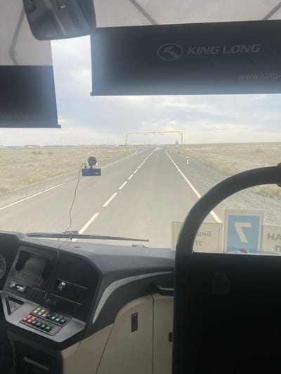 The image is taken from the interior of a bus, looking forward through the windshield. The road ahead is a two-lane asphalt road stretching into a flat, sparsely vegetated landscape under a cloudy sky. On the right side of the road, there are signs. A yellow gantry with overhead lights spans the road in the distance. A dashcam is mounted on the windshield. The dashboard of the bus is visible in the foreground, with various buttons and a screen. The exterior landscape appears to be arid, with some industrial structures visible in the far distance on the left. The location is identified as Қосшағыл ауылдық округі, Kazakhstan.