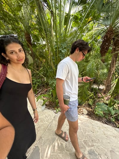 A young woman with long, dark hair is walking on a stone path in a tropical environment. She is wearing a black sleeveless dress and sunglasses. She has a thin gold necklace on. A young man, also with dark hair, is walking in front of her on the path. He is wearing a white t-shirt, blue and white striped shorts, sandals, and eyeglasses. He is looking at his phone. The environment is lush with green foliage. This scene is located on a path in the tropical forest of the Santa Teresa region, Costa Rica, at the approximate coordinates of 9.635 latitude, -85.161 longitude.