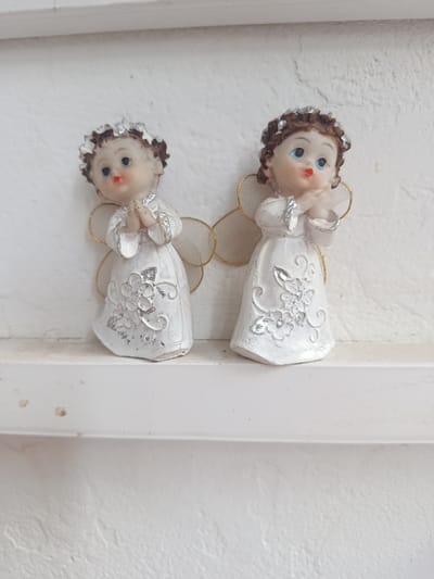 This media file features two small, decorative angel figurines positioned on a white shelf. The figurines are of cherubic figures, each with light skin, brown curly hair adorned with a silver headband, and wide blue eyes. They are dressed in flowing white gowns decorated with silver floral patterns and subtle glitter accents. Their hands are clasped together in front of their faces, suggesting a pose of prayer or gentle contemplation. Delicate, translucent wings with gold trim extend from their backs.
The figurines are placed against a textured white background, likely a wall, which appears slightly rough and uneven. The shelf itself is also white and shows some minor signs of wear or dust, with a few small brown spots visible on its surface. The lighting is bright and even, suggesting it might be daytime, possibly indoors in a well-lit room. There is no visible text in the image. The overall impression is one of innocence and peacefulness, commonly associated with angel motifs. The location context of Cupcini, Moldova, does not provide specific visual cues within the image itself.