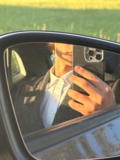 A close-up, reflected image shows a person taking a selfie in a car's side-view mirror. The person, wearing a white collared shirt and a gray jacket, is holding a black smartphone with a triple-lens camera. The background of the reflection reveals the interior of a car, with a window and dashboard visible. Outside the car, a blurred field of green grass and yellow highlights suggests a rural or park-like setting. The scene appears to be captured during daylight, with warm sunlight illuminating the person's face and hand. The image is framed by the black edge of the side-view mirror. No specific location in Нёманскі сельскі Савет, Belarus, is identifiable.