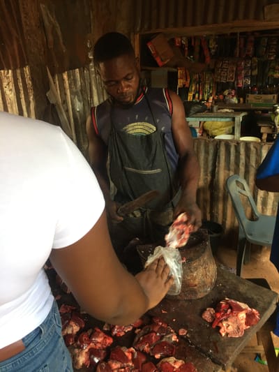 This media file captures a scene inside a market stall or roadside shop in Isieke, Nigeria. The primary focus is on a dark-skinned man, likely a butcher or vendor, who is preparing raw meat. He wears a dark apron over a t-shirt and has short dark hair, his gaze fixed on the task at hand. In his right hand, he holds a piece of raw meat, actively placing it into a clear plastic bag. Another person's hand, presumably a customer, holds the plastic bag in the foreground, ready to receive the purchase.
On a large, dark wooden chopping block, numerous pieces of raw red meat, resembling organ meats like liver, are visible. Adjacent to this main chopping surface is a heavy, cylindrical wooden log, likely used for additional butchering. The man also holds a large knife or cleaver, indicating his trade.
The setting is characterized by walls made of corrugated metal sheets, suggesting a simple, functional structure. In the background, shelves are densely packed with various packaged goods, including snacks and other items in colorful wrappers, implying a general store or a multi-purpose market stall. A blue plastic chair is partially visible in the upper background. The lighting is somewhat dim, typical of an indoor or sheltered market space. A person's arm and leg, wearing a white shirt and blue jeans, are prominent in the foreground, further indicating the presence of a customer. No discernible text is visible on any of the products or elements in the image. The scene portrays a routine commercial activity in a local market environment.
