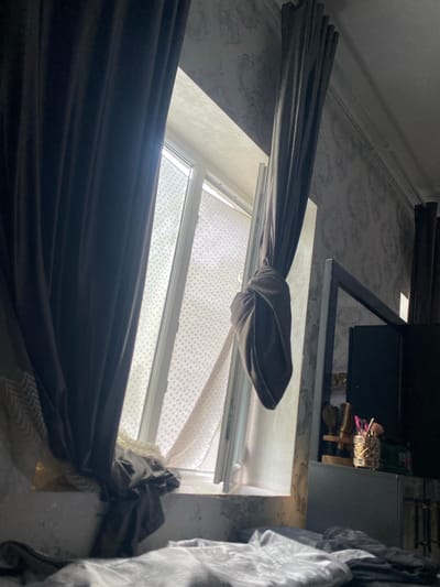 The image depicts an indoor scene, likely a bedroom, with natural light entering through a window covered by dark grey curtains. The curtains are partially drawn, with one side tied back, revealing a lighter, patterned sheer curtain behind it. The window appears to be in an urban setting, possibly in Боғистон, Tajikistan, judging by the architecture visible through the window and the style of the wallpaper. The room is furnished with what appears to be a bed in the foreground, partially covered with bedding. To the right of the window, a dark piece of furniture, possibly a dresser or cabinet, is visible. On top of this furniture, there are some decorative items, including what looks like a wooden stand holding brushes and a decorative gold container filled with pink-tipped items, possibly makeup brushes or pens. The wallpaper on the wall is patterned in a light grey floral or damask design. The overall atmosphere is dim, with shadows cast by the curtains and the angle of the sunlight. There are no people visible in the image.