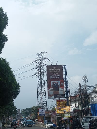 The image captures a bustling street scene under an overcast or hazy sky. A large, green, dense tree canopy occupies the left side of the frame, partially obscuring the sky and extending down towards the street level.
Dominating the central and upper-left portion of the image is a massive metal lattice high-voltage power transmission tower (pylon), from which several thick power lines extend horizontally across the sky. To the right of this power tower, a tall, prominent billboard stands vertically. The billboard features a dark background, likely red or brown, and advertises "GUDANG GARAM International FILTER" at the top. Below this, the text reads "KEMANTAPAN SANG RASA LEGENDARIS" (meaning "The Steadiness of the Legendary Taste" or "The Stability of the Legendary Flavor"). An image of a man in a suit, seated in a chair, is visible on the lower part of this billboard. Smaller text "PHA" and "BELL & BATO" are also present on the billboard.
Further to the right, a cellular communication tower with multiple antennas is visible, surrounded by a dense tangle of utility wires and poles, indicating an intricate urban infrastructure.
At street level, the road appears to be a multi-lane thoroughfare with a moderate amount of traffic. Numerous vehicles, including cars and a significant number of motorcycles, are present. Several individuals on motorcycles are visible, some wearing helmets. A white van is seen on the far right.
Along the right side of the road, various commercial establishments or buildings line the street, adorned with a variety of smaller signs and banners. One blue sign clearly displays "PUSAT GADAI" (meaning "Pawn Center"). Another yellow sign, partially obscured, shows "ZHAKY CELL" multiple times. People are visible near these establishments and on the street. The overall impression is a lively urban or suburban environment with continuous commercial activity and traffic flow during daytime. The sky's appearance suggests a cloudy day or possibly some atmospheric haze.