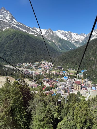 An aerial view looks down on the city of Dombay, Russia, nestled within a valley surrounded by the snow-capped Caucasus Mountains. The image captures a vibrant ski resort town with a dense forest of coniferous trees covering the slopes leading up to the majestic, rocky peaks. The town itself is a collection of colorful buildings, hotels, and residential structures, with a prominent cable car system weaving through the scene, its cables stretching across the frame and two gondolas visible in motion. The clear blue sky suggests fair weather.