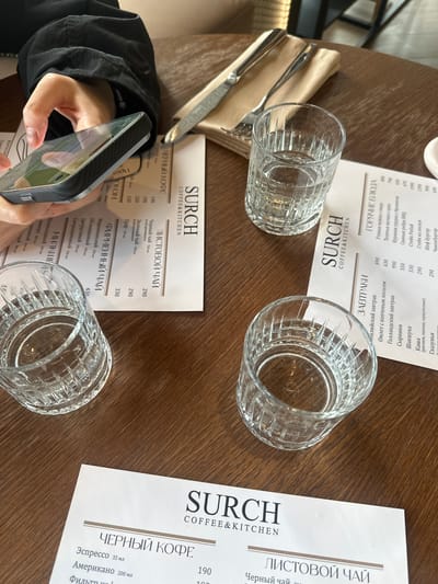 The image depicts a cafe or restaurant setting in Усады, Russia, viewed from an overhead perspective. A dark-grained wooden table occupies the majority of the frame. In the upper left, a person's left hand, clad in a dark, long-sleeved garment, holds a dark-cased smartphone horizontally. The phone screen displays reflections of the surrounding environment.
Three white menus, each bearing the logo "SURCH COFFEE & KITCHEN" at the top, are arranged on the table. They list various food and drink items in Russian, categorized under "ЧЕРНЫЙ КОФЕ" (Black Coffee), "ЛИСТОВОЙ ЧАЙ" (Loose Leaf Tea), "ФИРМЕННЫЙ ЧАЙ" (Signature Tea), "ЗАВТРАКИ" (Breakfasts), and "ГОРЯЧИЕ БЛЮДА" (Hot Dishes), with corresponding prices.
Three clear, empty glasses with a prominent vertical fluted design are distributed across the table. One glass in the upper center is partially filled with water, another in the lower left is also partially filled, and a third in the lower center appears empty. In the upper central area, a metal knife and fork rest on a folded beige napkin. The background is blurred, showing indistinct elements of the establishment's interior.