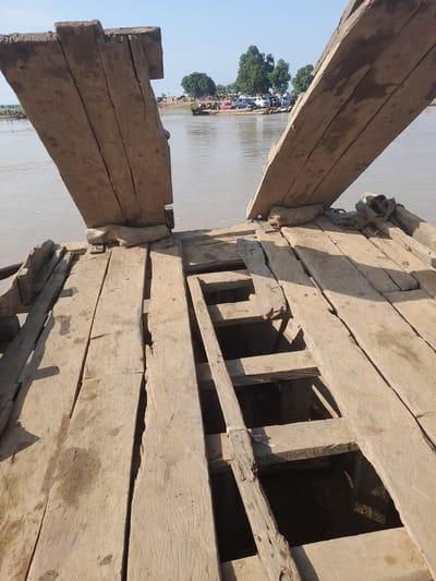 The image depicts a ferry crossing a river in Nemnei, Nigeria. The foreground is dominated by the weathered wooden deck of a ferry, revealing gaps and structural supports. Two large, angled wooden planks extend upwards from the ferry deck, forming a makeshift ramp or entryway. In the background, the murky river flows towards a far bank lined with trees and a small settlement. Several other ferries are visible on the water, some laden with vehicles, including cars and what appear to be trucks. Numerous people are gathered on the ferries and the riverbank, suggesting an active transportation hub. The overall scene conveys a sense of rudimentary but functional riverine transport.