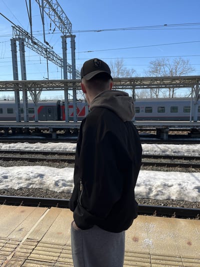 A person wearing a black jacket, gray hoodie, and baseball cap stands on a metal platform at a railway station.  Tracks with snow bordering them are visible.  Two passenger trains are stationed at the platform.  Overhead electrical structures and wires are present.  The sky is blue with a few bare trees in the background.  The location is likely a railway station in Tashevka, Russia.
