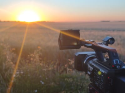 A semi-circular sunrise with bright yellow rays illuminates a foggy, grassy field. In the foreground, a black Sony video camera on a tripod is pointed towards the sunrise. The camera has a lens, a handle, and visible buttons and indicators. The environment appears rural and possibly damp due to the fog. The image has a shallow depth of field, with the background and foreground being out of focus. The scene is likely set in Tyulenovo, Bulgaria, given the context provided.