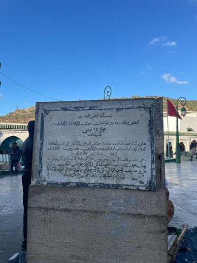 The image depicts a public courtyard area within the city of Moulay Idriss Zerhoun, Morocco, specifically near the Mausoleum of Moulay Idriss I. The central subject is a weathered, rectangular commemorative plaque, primarily white with black Arabic script, affixed to a multi-layered grey stone base. The inscription on the plaque identifies it as related to Moulay Idriss Al-Akbar and King Mohammed VI, dated 20 Jumada al-Akhira 1425 AH, corresponding to October 6, 2004 AD.
To the left of the plaque, an individual in a dark hooded jacket stands facing away from the camera, partially obscured by the plaque. Further left, a second individual wearing a cap and jacket is visible near an arched entrance of a light-colored building featuring green-tiled roof accents. In the background to the right of the plaque, a third, indistinct individual is present near another structure.
The environment includes a paved, light-colored courtyard. In the mid-ground right, a tall pole supports a red Moroccan flag and a traditional green lantern. Beyond the structures, rolling hills with sparse vegetation are visible under a clear, bright blue sky with minimal cloud cover. Partially visible on the ground to the lower right of the plaque is a stringed musical instrument, possibly a _guembri_, resting on a blue fabric. No direct interactions between individuals are discernible.