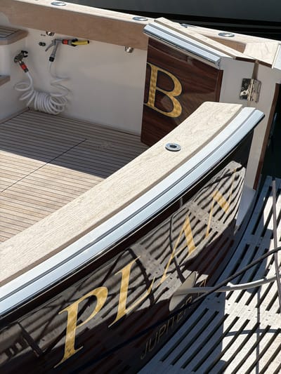 This image shows the stern of a wooden boat docked in the Village of Sag Harbor, United States of America. The boat features a polished wooden deck and a dark, reflective hull with gold lettering that spells out "PLANET" and "JUPITER." A coiled white hose with a nozzle is visible on the port side, attached to the white interior wall of the boat. A stainless steel fitting is visible on the deck. The boat is docked next to a wooden pier. The scene is illuminated by bright sunlight, casting shadows on the deck and hull.