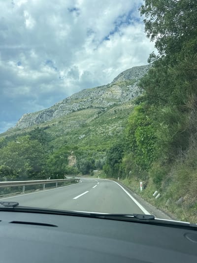 This image captures a scenic drive along a winding road in Rijeka Reževići, Montenegro. The perspective is from inside a vehicle, looking out towards a dramatic mountainous landscape. 

The main subjects are the imposing, rocky mountainside covered in lush green vegetation and the asphalt road curving to the right. The road has white lane markings, and a metal guardrail runs along its edge. Trees and bushes line the roadside, with denser foliage on the upper slopes of the mountain. In the distance, the road disappears around a bend, suggesting a journey through a mountainous region. The sky is partly cloudy, with patches of blue sky visible amongst the white and grey clouds. 

The scene conveys a sense of travel and exploration, with the natural beauty of the Montenegrin coast being a prominent feature. The time of day appears to be daytime, given the natural light and the visible clouds. The weather seems to be mild, with a mix of sun and clouds. There are no people or specific objects of interest, other than the road infrastructure and the natural elements. No visible text is present in the image.