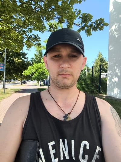 This is a selfie of a man in his late 20s or early 30s, taken outdoors on a sunny day. He is wearing a black baseball cap, a black tank top with white lettering, and a necklace with a cross pendant. He has short brown hair and stubble. His right arm is visible, showing a tattoo. The background suggests a street or sidewalk next to a road with trees and buildings. The lighting indicates it's daytime. The man's expression is neutral. The white lettering on his tank top appears to spell out "LENIC", possibly part of a larger word or phrase. The image was taken in Wolgast, Germany.
